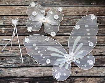 May include: A set of fairy wings and a wand. The wings are white, with a sheer fabric and floral accents. The wand is white with a flower on top. The items are arranged on a wooden surface.