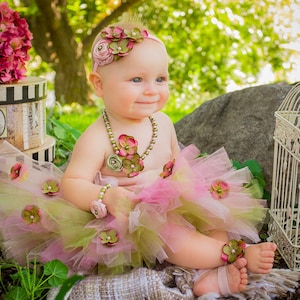 May include: A baby girl wearing a pink and green tutu with flower embellishments, a flower headband, and a pearl necklace. She is sitting on a blanket with a birdcage and a box in the background.