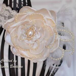 Puede incluir: Una flor de sat&eacute;n blanco con detalles de perlas y un centro de pedrer&iacute;a dorada. La flor est&aacute; unida a una diadema blanca con plumas y perlas.