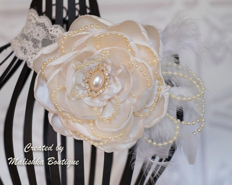 Puede incluir: Una flor de sat&eacute;n blanco con detalles de perlas y un centro de pedrer&iacute;a dorada. La flor est&aacute; unida a una diadema blanca con plumas y perlas.
