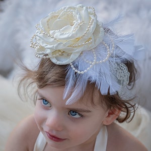 Puede incluir: Una diadema blanca con una flor, detalles de perlas y plumas blancas. La diadema la lleva una ni&ntilde;a con el pelo casta&ntilde;o y los ojos azules.