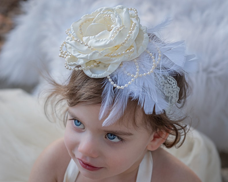 Puede incluir: Una diadema blanca con una flor, detalles de perlas y plumas blancas. La diadema la lleva una ni&ntilde;a con el pelo casta&ntilde;o y los ojos azules.