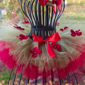 May include: A layered tutu skirt in gold, pink, and red tulle, adorned with red flower accents and a red satin bow. The tutu is displayed on a black wire dress form. The text "Created by Malishka Boutique" is visible.