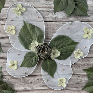 May include: White fairy wings with green leaves and white flowers. The wings are decorated with silver glitter and small beads.