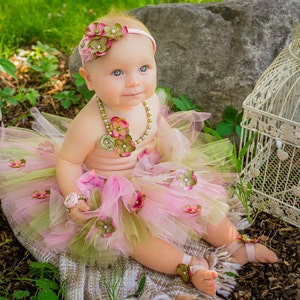 Puede incluir: Una ni&ntilde;a peque&ntilde;a con un tut&uacute; rosa y verde con adornos de flores, una diadema de flores y un collar de perlas. Est&aacute; sentada en una manta marr&oacute;n y blanca con una jaula de p&aacute;jaros en el fondo.