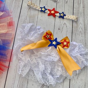 May include: White lace ruffle bloomers with a gold ribbon and red, blue, and gold star embellishments. A headband with red, blue, and gold stars is also included.