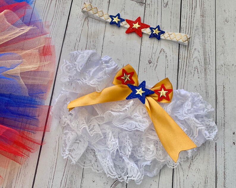 May include: White lace ruffle bloomers with a gold ribbon and red, blue, and gold star embellishments. A headband with red, blue, and gold stars is also included.