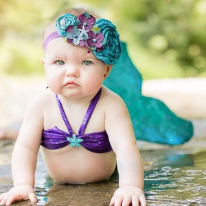 May include: A baby wearing a purple and teal mermaid costume with a flower headband. The baby is crawling on a rock near a stream of water.