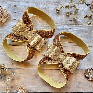 Op de afbeelding: Twee gouden glitter haarbandjes met een grote gouden glitter strik op elk. De haarbandjes liggen op een houten oppervlak.