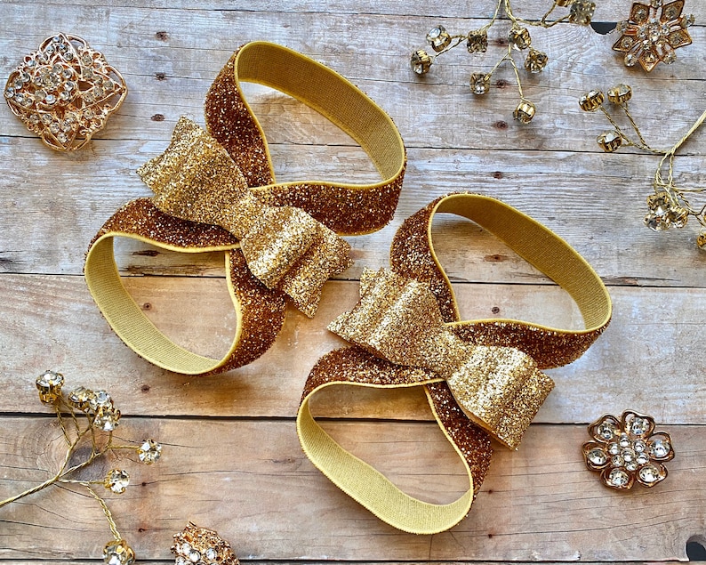 Op de afbeelding: Twee gouden glitter haarbandjes met een grote gouden glitter strik op elk. De haarbandjes liggen op een houten oppervlak.