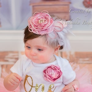 May include: A baby girl wearing a white onesie with the word "One" in gold glitter, a pink flower embellishment, and a pink and white headband with a feather. The baby is sitting on a pink tulle skirt.