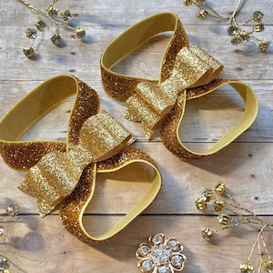 Op de afbeelding: Twee gouden glitter haarbandjes met grote gouden glitter strikken. De haarbandjes liggen op een houten oppervlak.