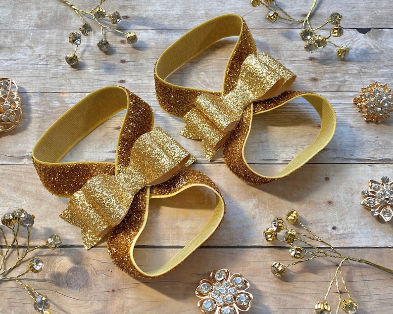 Op de afbeelding: Twee gouden glitter haarbandjes met grote gouden glitter strikken. De haarbandjes liggen op een houten oppervlak.