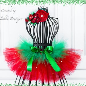 May include: Red and green tulle tutu skirt with a green satin bow and white pearl accents. The skirt is displayed on a black mannequin. The image also includes a red flower headband with white pearl accents.