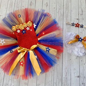 May include: A red, white, and blue tulle tutu with gold glitter and stars. The tutu has a red crocheted bodice with gold stars and a gold bow. The set also includes a white lace headband with a gold bow and a white lace diaper cover with a gold bow.