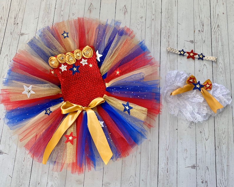 May include: A red, white, and blue tulle tutu with gold glitter and stars. The tutu has a red crocheted bodice with gold stars and a gold bow. The set also includes a white lace headband with a gold bow and a white lace diaper cover with a gold bow.