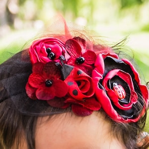 May include: A black headband with red and black fabric flowers and a ladybug accent. The headband is decorated with black polka dots and a black feather.