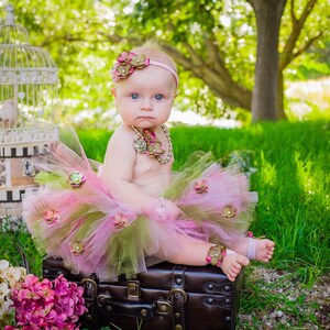 Puede incluir: Una ni&ntilde;a peque&ntilde;a con un tut&uacute; rosa y verde con detalles de flores est&aacute; sentada en una maleta vintage. Lleva una diadema de flores y un collar de oro.