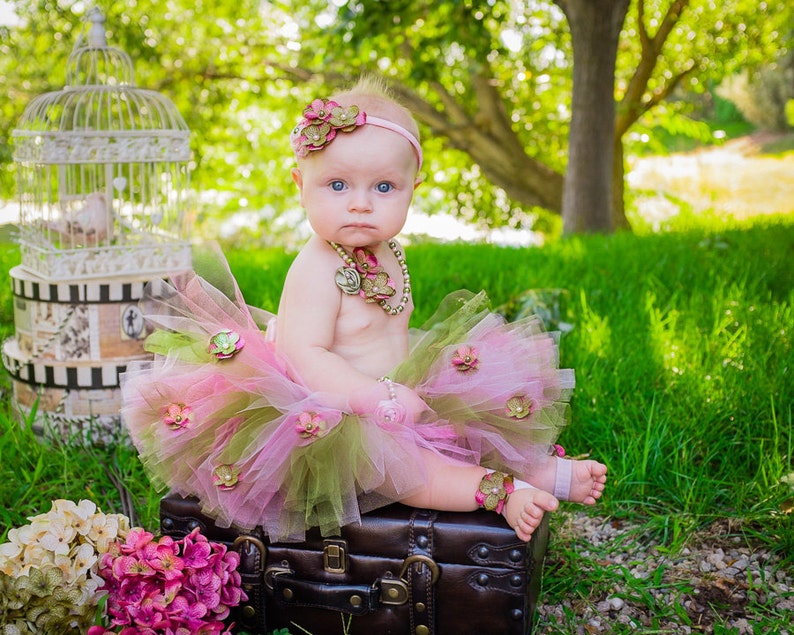 Puede incluir: Una ni&ntilde;a peque&ntilde;a con un tut&uacute; rosa y verde con detalles de flores est&aacute; sentada en una maleta vintage. Lleva una diadema de flores y un collar de oro.