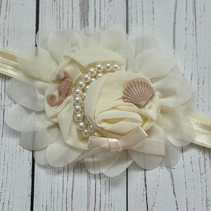 May include: A white headband with a large, ivory flower made of fabric. The flower is decorated with a pearl strand, a small seashell, and a seahorse charm.