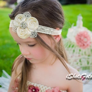 May include: A young girl with long brown hair wearing a white lace headband with a large ivory satin flower and a rhinestone embellishment. She is wearing a white tulle dress with a pink and ivory flower detail.