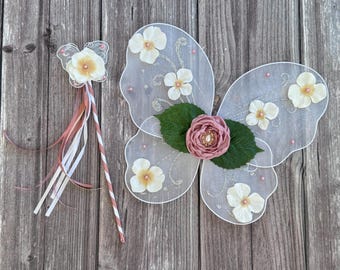 May include: White fairy wings with a pink flower and green leaves. The wings are decorated with glitter and small white flowers. A white butterfly wand with pink and white ribbons is also included.