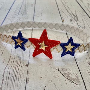May include: A headband with a white and gold patterned band and three stars. The stars are red, blue, and gold.