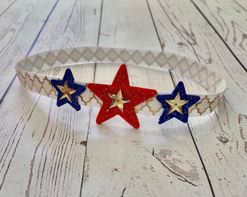 May include: A headband with a white and gold patterned band and three stars. The stars are red, blue, and gold.