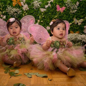 Puede incluir: Dos beb&eacute;s con trajes de hada rosa y verde con alas de mariposa. Los beb&eacute;s est&aacute;n sentados en un suelo de madera con hojas verdes esparcidas a su alrededor.