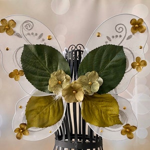 May include: White butterfly wings with gold glitter and green leaves. The wings are decorated with gold flowers and beads.