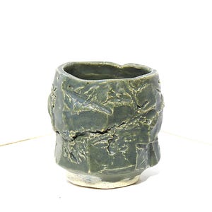 May include: A handmade, textured ceramic cup in a muted green-gray hue. The cup has a rough, uneven surface with a light-colored base. The design gives the cup a rustic, organic appearance, suitable for beverages or as a decorative piece.