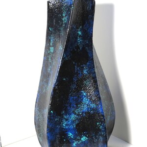 May include: A tall, abstract ceramic vase with a textured surface. The vase features a dark blue and black color scheme with splashes of turquoise. The form is geometric and twisted, creating a modern aesthetic. The vase is displayed against a white background.