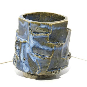 May include: A handcrafted ceramic cup with a unique, textured design. The cup features a hexagonal top and a rough, layered exterior with shades of blue and brown. The base is a lighter, unglazed color. This artful piece is ideal for home decor.