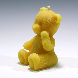 May include: A yellow teddy bear candle, sitting with its paw near its face. The candle has a textured surface and a wick at the top of its head. The bear is made of wax and has a classic teddy bear shape.