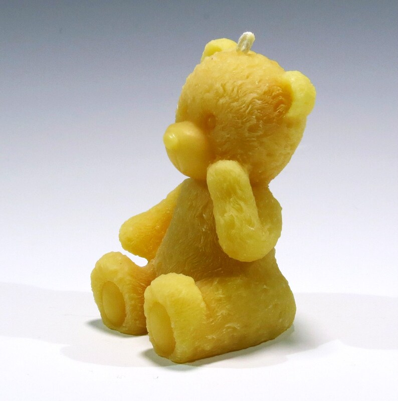 May include: A yellow teddy bear candle, sitting with its paw near its face. The candle has a textured surface and a wick at the top of its head. The bear is made of wax and has a classic teddy bear shape.