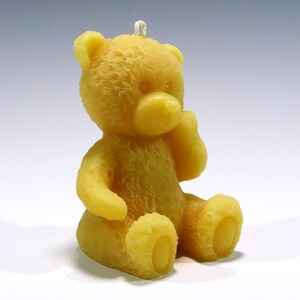 May include: A yellow teddy bear candle with a sitting pose. The candle has a textured surface and a loop at the top for hanging. The bear's features include a nose, ears, arms, and feet. The candle is made of wax.