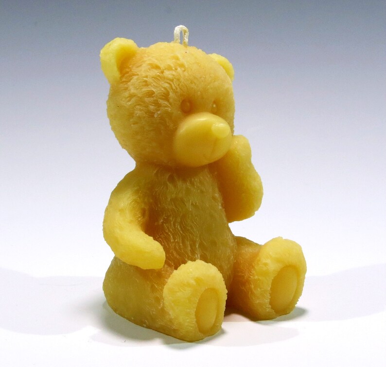 May include: A yellow teddy bear candle with a sitting pose. The candle has a textured surface and a loop at the top for hanging. The bear's features include a nose, ears, arms, and feet. The candle is made of wax.