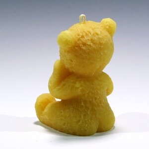 May include: A yellow teddy bear candle, sitting with its hands clasped. The candle has a textured surface and a loop at the top for hanging. The bear is in a seated position, with its legs tucked in. The candle is a solid yellow color.