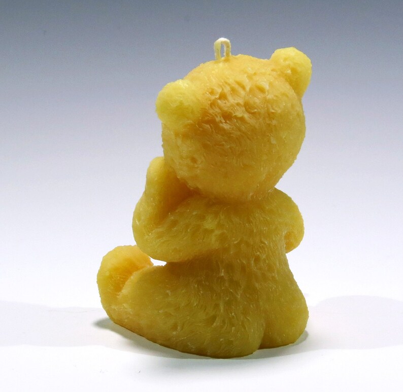 May include: A yellow teddy bear candle, sitting with its hands clasped. The candle has a textured surface and a loop at the top for hanging. The bear is in a seated position, with its legs tucked in. The candle is a solid yellow color.