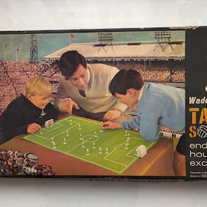 Könnte beinhalten: Eine Vintage-Spieleschachtel von Waddington's Table Soccer mit einer Illustration von zwei Kindern und einem Erwachsenen, die auf einem grünen Spielfeld spielen. Die schwarze Seitenwand zeigt eine weiße Spielfigur und den Text Waddington's TABLE SOCCER endless hours of excitement.