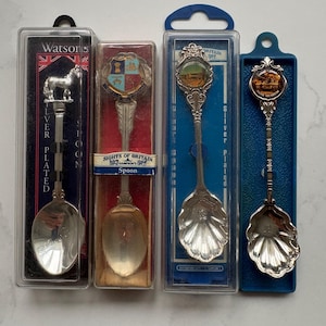Könnte beinhalten: Vier versilberte Souvenir-Löffel in einzelnen Kunststoff-Präsentationsboxen. Die Löffel haben dekorative Griffe mit Emblemen, darunter ein Pferd, ein Wappen und kreisförmige Motive. Die Verpackung enthält Texte wie Watsons, Sights of Britain und Tank Museum sowie durchsichtige Kunststoffabdeckungen.