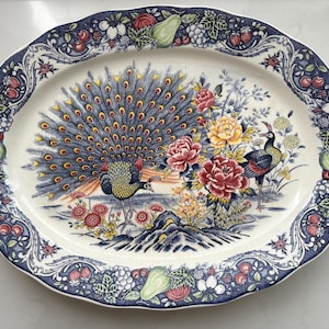 Op de afbeelding: Een ovale keramische serveerschaal met een centrale afbeelding van een pauw met een uitgespreide staart, een tweede vogel en kleurrijke bloemen. De geschulpte rand is versierd met een donkerblauwe boord met patronen van peren en druiven.