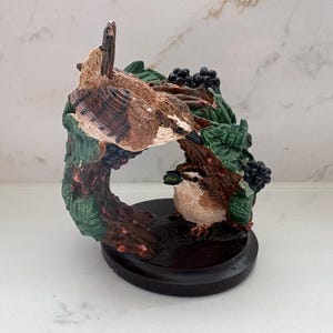 Könnte beinhalten: Eine dekorative Figur mit zwei kleinen braun-weißen Vögeln, die auf einem strukturierten Ast mit grünen Blättern und dunklen Beerenclustern sitzen. Die Vögel befinden sich in einer kreisförmigen Aststruktur auf einem runden schwarzen Sockel.