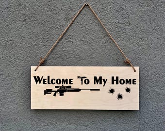 May include: Wooden sign with the text "Welcome To My Home" in black. The sign features a rifle and bullet hole graphics. It is suspended by a natural fiber rope.