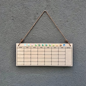 Puede incluir: Un calendario de pared de madera con una cuadrícula para la planificación semanal, suspendido por una cuerda natural. El calendario tiene iconos decorativos encima de cada día de la semana. El fondo es una pared gris texturizada.