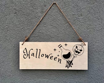 May include: A rectangular wooden sign with the word "Halloween" in a decorative font. The sign features a potion bottle, stars, and a jack-o'-lantern lollipop design. It hangs from a jute rope against a textured gray wall.