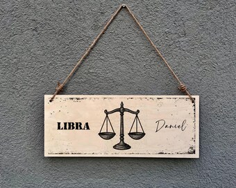 May include: Wooden Libra sign with a distressed border, featuring the word "LIBRA" and the name "Daniel". A scale graphic is centered on the sign. The sign hangs from a jute rope.