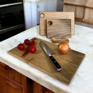 Puede incluir: Una tabla de cortar de madera grande con un cuchillo, una cebolla, un pimiento rojo y tomates. La tabla tiene un agujero en la esquina superior y está hecha de madera oscura. La tabla de cortar está sobre una encimera blanca.