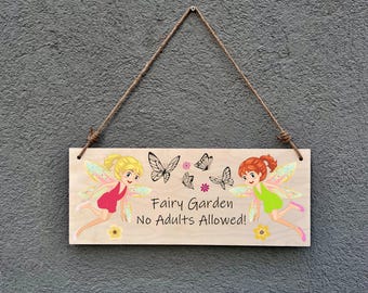 May include: Wooden sign with the text "Fairy Garden No Adults Allowed!" featuring illustrations of fairies and butterflies. The sign is rectangular, with a natural wood grain finish, and is suspended by a brown rope.