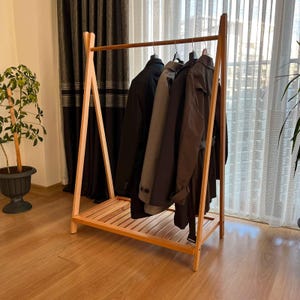 Op de afbeelding: Een lichtgekleurd houten A-vormig kledingrek met een horizontale roede en een onderste plank met latten. Vier jassen in de kleuren zwart, grijs en donkerbruin hangen aan individuele kledinghangers netjes op een rij aan de roede.